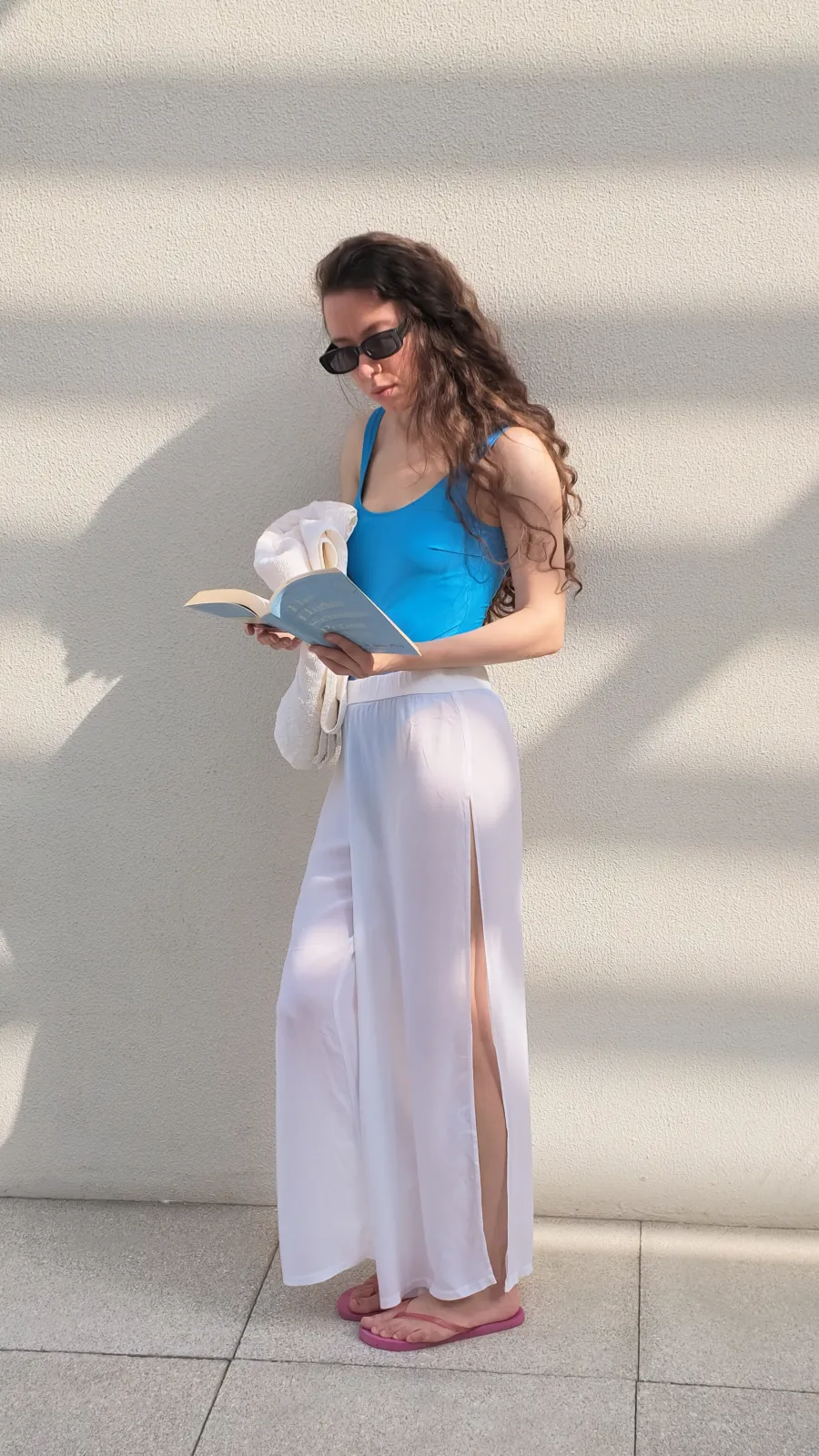 Olga in a blue swimsuit reading a book with a towel on her shoulder, sunlit wall at the Emaar South villa pool
